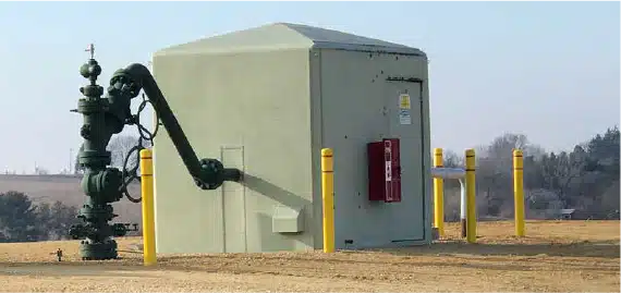 A small utility enclosure with a large pipe connected to equipment outside, surrounded by yellow safety bollards, stands in an open, grassy area with trees in the background.